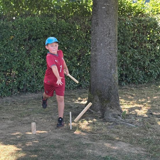 Ein Junge in einem roten Trikot mit der Nummer 7 spielt Cricket mit einem Schläger in der Nähe eines Baumes in einem Garten.