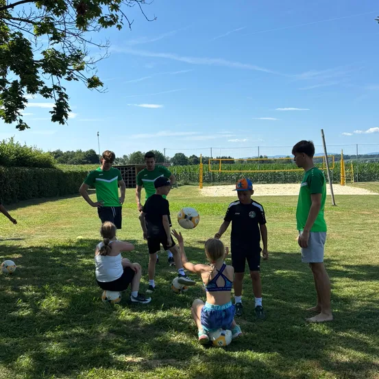 Eine Gruppe von Menschen spielt Fußball in einem offenen Bereich mit einem Volleyballnetz und Bäumen. Einige knien und halten Fußballbälle.