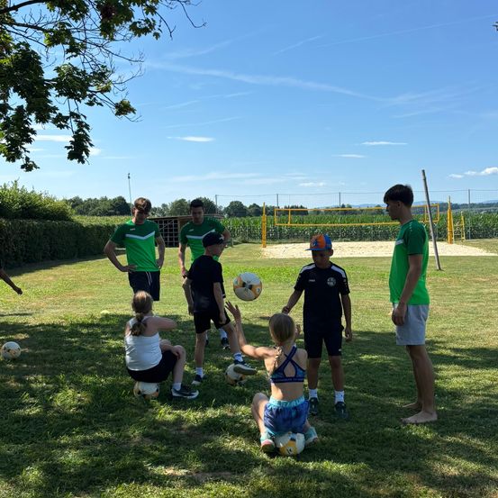Eine Gruppe von Menschen spielt Fußball in einem offenen Bereich mit einem Volleyballnetz und Bäumen. Einige knien und halten Fußballbälle.