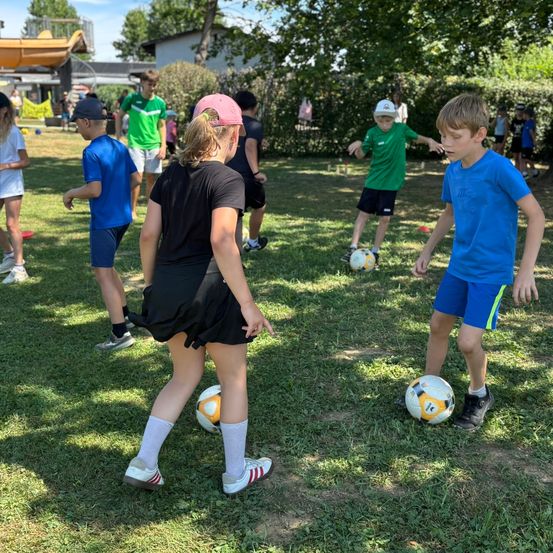 Eine Gruppe von Kindern spielt Fußball auf einem Rasenfeld, mit einer Frau vor ihnen und einem Jungen in Blau dahinter. Sie üben mit gelben und weißen Fußballen.