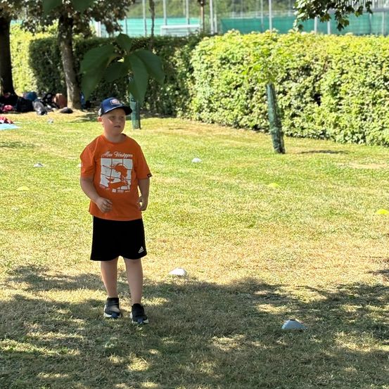 Ein junger Junge in einem orangefarbenen T-Shirt und einer Baseballkappe steht in einem Grasbereich mit einigen Bäumen und Büschen.