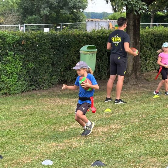 Ein Junge rennt mit einem Football auf einem Rasenfeld, umgeben von Trainingshütchen. Ein Mann hält einen Football hinter ihm. Ein Mädchen steht im Hintergrund in der Nähe eines grünen Mülleimers.