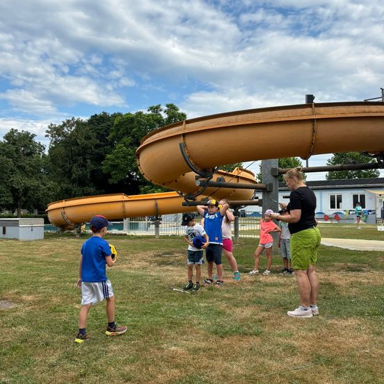 Eine Gruppe von Kindern und Erwachsenen, einschließlich einer Frau, steht in der Nähe eines Wasserrutsches. Einige Kinder halten Baseballhandschuhe. Der Rutsch ist gelb und hat eine gebogene Form. Im Hintergrund sind Bäume und ein Gebäude zu sehen.