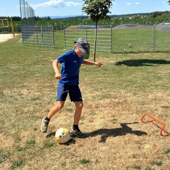 Ein Junge in Blau rennt auf einen Fußball auf einem Rasenfeld zu, mit einem Baum und Zäunen im Hintergrund.