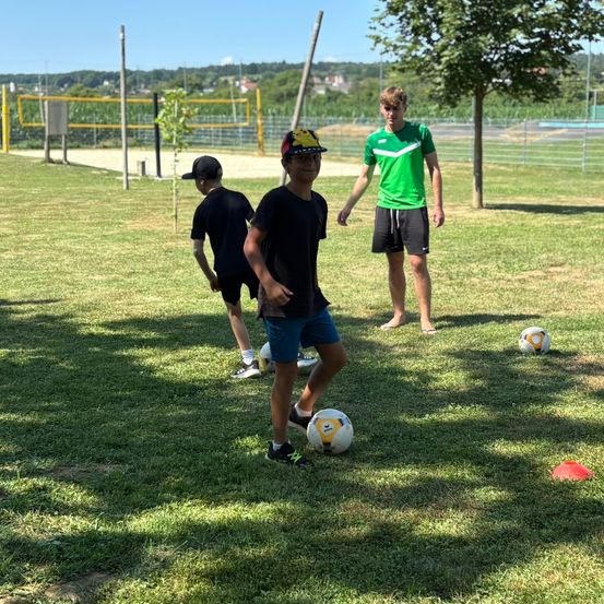 Drei Jungen üben Fußball auf einem Rasenfeld. Einer trägt einen Hut, ein anderer hat einen Fußball. Dahinter beobachtet ein Trainer.