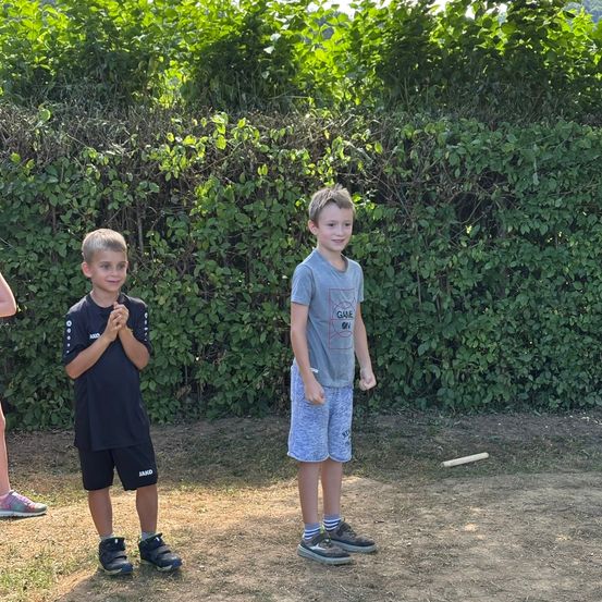 Zwei Jungen stehen vor einer Hecke, einer lächelt und der andere hat die Hände gefaltet. Ein Stock liegt vor ihnen auf dem Boden.