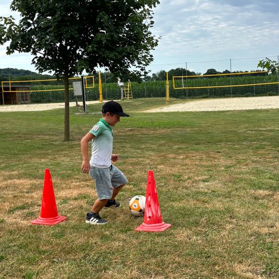 Ein junger Junge übt Fußballübungen auf einem Feld und nutzt Kegel als Wegweiser. Ein Fußball liegt in der Nähe, und er trägt Turnschuhe. Im Hintergrund befinden sich ein Baum und ein Volleyballnetz.