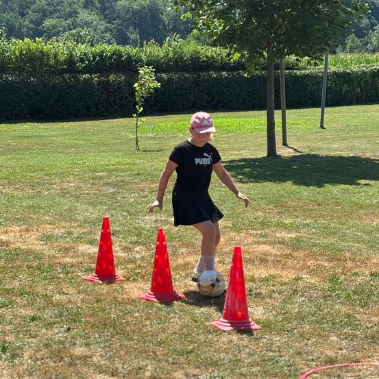 Ein Mädchen in einem schwarzen Puma-Shirt und Hut übt Fußball auf einem Feld mit Kegeln und einem Ball.