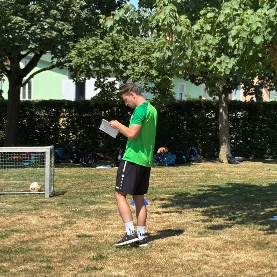 Ein Mann in einem grünen T-Shirt und schwarzen Shorts steht in einem Park und liest ein Blatt Papier. In der Nähe sind ein Fußballtor, Bäume und Gebäude zu sehen.