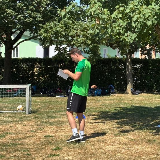 Ein Mann in einem grünen T-Shirt und schwarzen Shorts steht in einem Park und liest ein Blatt Papier. In der Nähe sind ein Fußballtor, Bäume und Gebäude zu sehen.