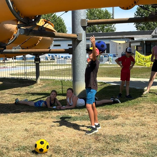Ein Junge hält einen Fußball und bereitet sich darauf vor, ihn auf einem Outdoor-Spielplatz zu schießen, wo drei Kinder im Hintergrund auf dem Gras liegen.