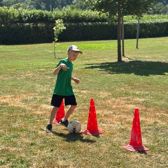 Ein junger Junge in einem grünen Shirt übt das Kicken eines Fußballs zwischen roten Kegel auf einem Grasfeld.