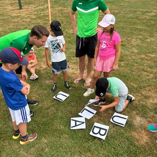 Mehrere Kinder und Erwachsene spielen ein Spiel auf einem Grasfeld. Buchstaben liegen auf dem Boden, und Kinder heben sie auf. Ein Kind kniet, während es einen Buchstaben aufhebt.