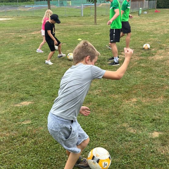 Ein junger Junge übt Fußball mit einem Ball auf einem Rasenfeld. Mehrere andere Kinder und Erwachsene sind ebenfalls anwesend.