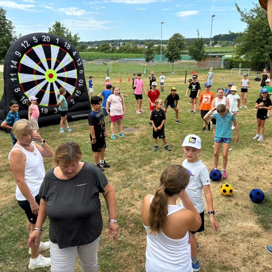 Eine Gruppe von Kindern und Erwachsenen spielt ein Spiel auf einem Grasfeld mit einem großen aufblasbaren Dartboard.