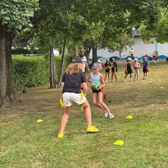 Eine Gruppe von Kindern spielt Rugby mit einem Trainer unter Bäumen. Einige Erwachsene beobachten.