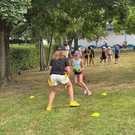 Eine Gruppe von Kindern spielt Rugby mit einem Trainer unter Bäumen. Einige Erwachsene beobachten.