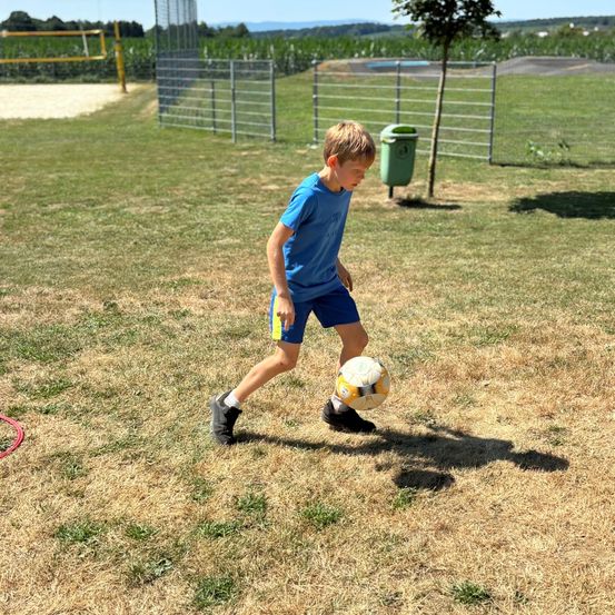 Ein junger Junge in einem blauen T-Shirt und Shorts übt das Kicken eines Fußballs auf einem offenen Feld. Hinter ihm befindet sich ein Zaun und ein grüner Mülleimer.