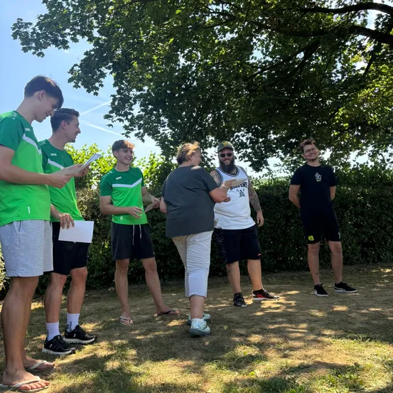 Sechs junge Männer in grünen Trikots und eine Frau in Grau stehen auf einem Feld, ein bärtiger Mann in einem weißen T-Shirt dazwischen. Sie scheinen zu sprechen.