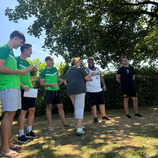 Sechs junge Männer in grünen Trikots und eine Frau in Grau stehen auf einem Feld, ein bärtiger Mann in einem weißen T-Shirt dazwischen. Sie scheinen zu sprechen.