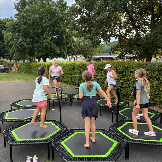 Eine Gruppe junger Mädchen übt das Springen auf Trampolinen in einem Außenbereich. Im Hintergrund befinden sich Bäume und Büsche.