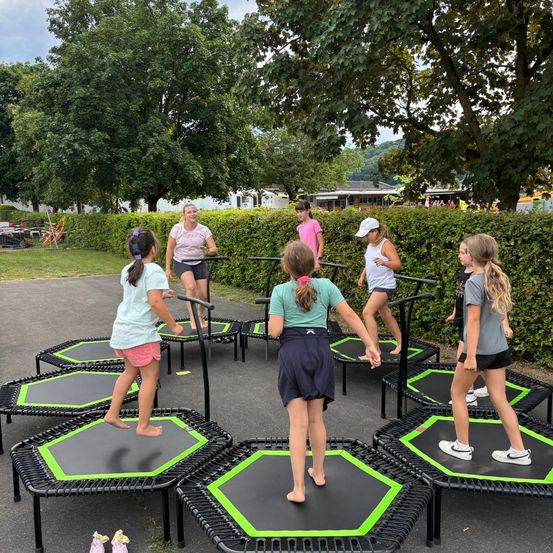 Eine Gruppe junger Mädchen übt das Springen auf Trampolinen in einem Außenbereich. Im Hintergrund befinden sich Bäume und Büsche.