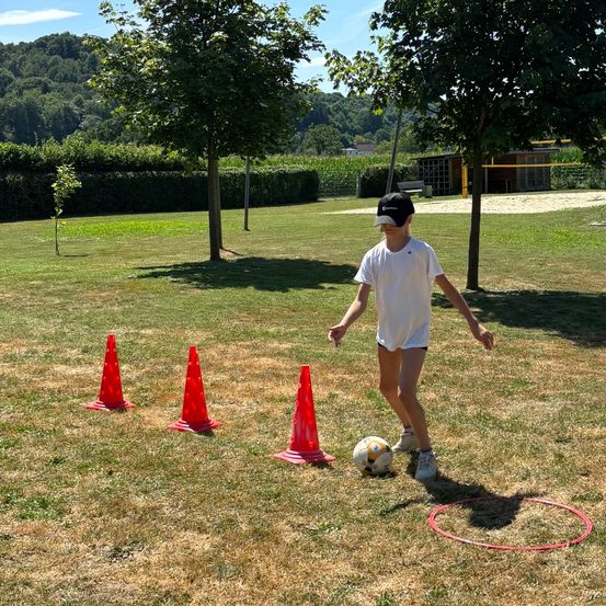 Ein junger Junge spielt Fußball, tritt einen Ball durch rote Kegel und zielt auf einen roten Hula-Hoop auf einem offenen Feld.