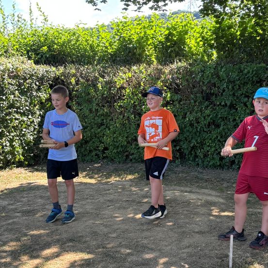 Drei Jungen stehen in einem offenen Bereich und halten Holzstöcke in der Hand, wahrscheinlich spielen sie ein Spiel. Der Junge auf der linken Seite trägt ein blaues Shirt und schwarze Shorts, der mittlere Junge ein orangefarbenes Shirt und schwarze Shorts und der Junge auf der rechten Seite ein rotes Shirt und rote Shorts.