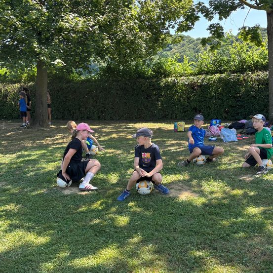 Eine Gruppe von Kindern sitzt auf dem Rasen, wahrscheinlich in einem Park, mit Fußballbällen. Ein Kind trägt eine Kappe und einen Hut. Einige tragen Turnschuhe. Im Hintergrund stehen einige Leute in der Nähe eines Baumes, andere sitzen auf dem Rasen.