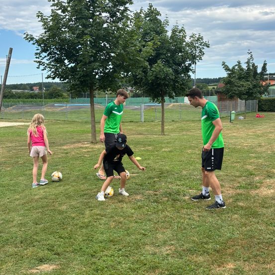 Zwei Männer in grünen Trikots beobachten einen Jungen und ein Mädchen in Pink und Schwarz, wie sie auf einem Rasenfeld unter Bäumen Fußballbälle treten.