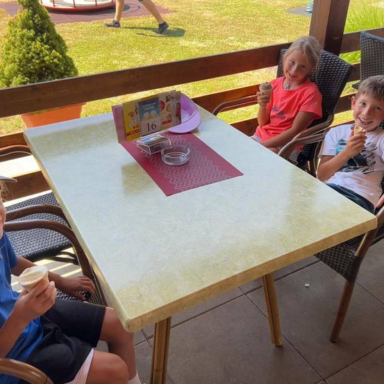 Drei Kinder genießen Eis am Außentisch, eines sitzt in einem Stuhl. Ein Topf und ein Spielplatz sind im Hintergrund zu sehen.