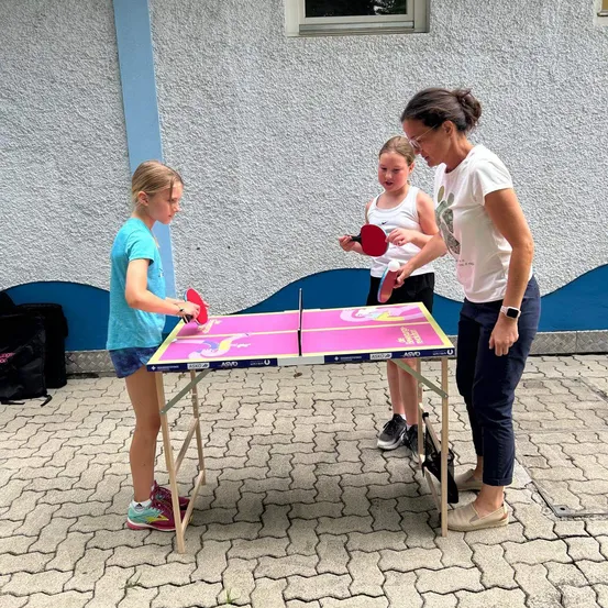 Drei Personen spielen Tischtennis auf einem tragbaren Tisch im Freien. Eine Frau gibt zwei jungen Mädchen Anweisungen.