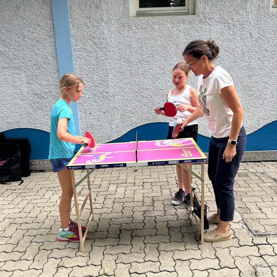 Drei Personen spielen Tischtennis auf einem tragbaren Tisch im Freien. Eine Frau gibt zwei jungen Mädchen Anweisungen.