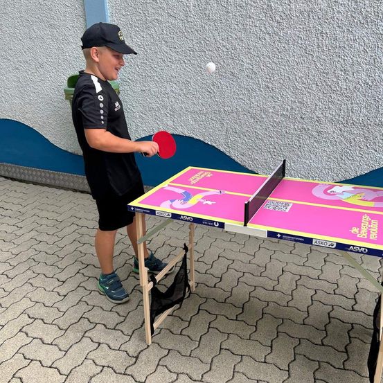 Ein Junge spielt draußen Tischtennis. Er trägt ein schwarzes Shirt und Shorts. Er hält eine rote Schläger und trifft den Ball. Der Tisch hat eine rosa Oberfläche mit Text und Logo.