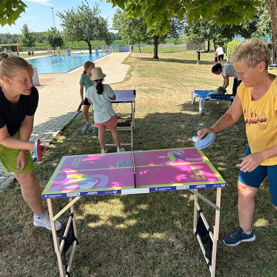 Mehrere Menschen spielen Tischtennis in einem Park. Eine Frau mit einem Schläger in der Hand ist kurz davor, den Ball zu schlagen. Eine andere Frau beobachtet sie. Dahinter befindet sich ein Schwimmbecken und weitere Menschen.