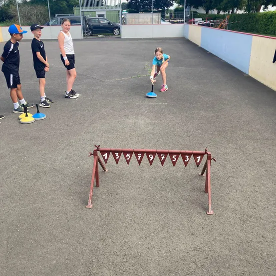 Vier Kinder spielen ein Spiel auf einem Betonplatz. Ein Mädchen beugt sich vor und spielt mit einem blauen Objekt. Drei Jungen stehen dahinter und beobachten.