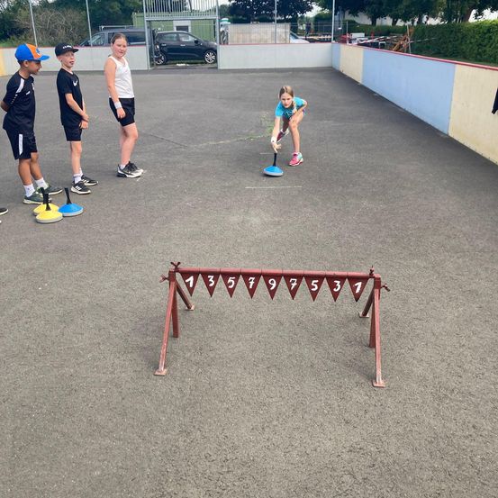 Vier Kinder spielen ein Spiel auf einem Betonplatz. Ein Mädchen beugt sich vor und spielt mit einem blauen Objekt. Drei Jungen stehen dahinter und beobachten.