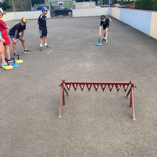 Kinder spielen ein Geschicklichkeitsspiel in einem Außenbereich mit einem roten Hindernisparcours. Sie tragen alle Turnschuhe und Mützen. Der Hindernisparcours hat Nummern darauf.