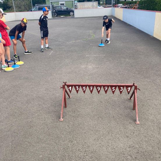Kinder spielen ein Geschicklichkeitsspiel in einem Außenbereich mit einem roten Hindernisparcours. Sie tragen alle Turnschuhe und Mützen. Der Hindernisparcours hat Nummern darauf.
