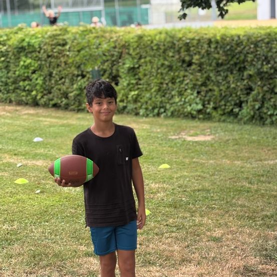 Ein Junge steht auf einem Rasenfeld, hält einen Football und hat einen Busch und andere Kinder im Hintergrund.