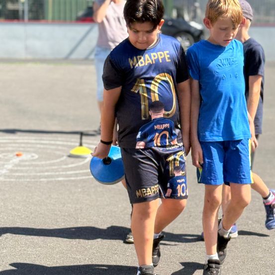 Ein Junge in einem Mbappe-Trikot geht neben einem anderen Jungen mit einem blauen T-Shirt. Sie tragen beide Shorts. Eine Person mit einem Armband ist hinter ihnen.