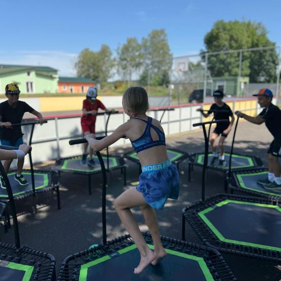 Kinder in Sportausrüstung springen im Freien auf Trampolins. Ein Mädchen in blauen Shorts springt in der Mitte. Im Hintergrund sind Bäume und ein Gebäude zu sehen.