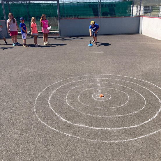 Ein Junge wirft einen Ball in ein Ziel auf einem gepflasterten Außenbereich, während mehrere Kinder zusehen.