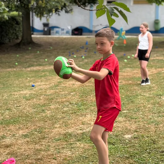 Ein Junge hält einen Fußball im Freien in einem Feld mit einem Mädchen im Hintergrund, umgeben von einem Gebäude und Bäumen.