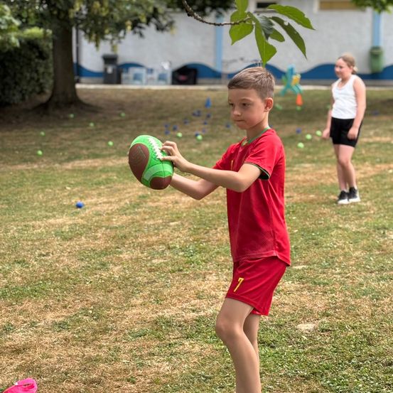 Ein Junge hält einen Fußball im Freien in einem Feld mit einem Mädchen im Hintergrund, umgeben von einem Gebäude und Bäumen.