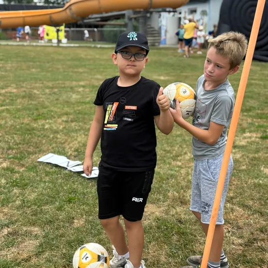 Zwei Jungen stehen auf einem Feld und halten Fußballbälle. Einer trägt ein schwarzes Shirt und eine Kappe, der andere ein graues Shirt. Im Hintergrund sind eine Rutsche und Luftballons zu sehen.