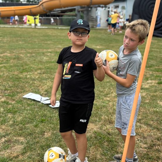 Zwei Jungen stehen auf einem Feld und halten Fußballbälle. Einer trägt ein schwarzes Shirt und eine Kappe, der andere ein graues Shirt. Im Hintergrund sind eine Rutsche und Luftballons zu sehen.