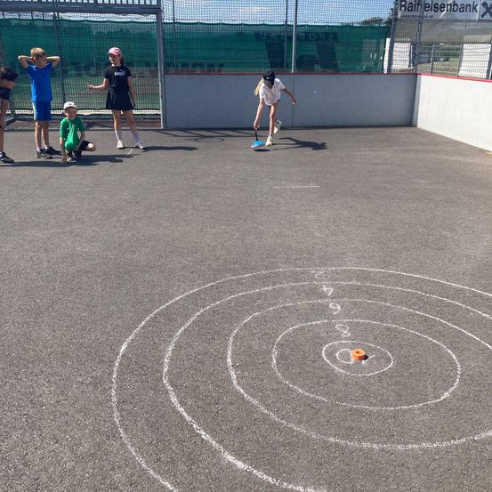 Eine Gruppe von Kindern spielt mit einem Frisbee auf einer Betonfläche. Sie stehen um ein Ziel mit einem Spiralmuster herum.