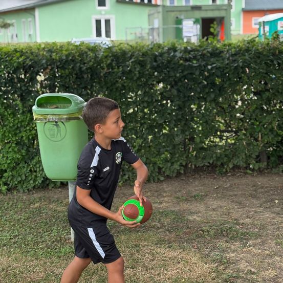 Ein Junge in einem schwarzen Sporttrikot hält einen Football in einem Park mit einem grünen Mülleimer im Hintergrund.