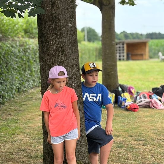 Zwei Kinder, ein Mädchen und ein Junge, stehen neben einem Baum in einem Grasfeld. Dahinter befinden sich Rucksäcke und eine Holzstruktur.